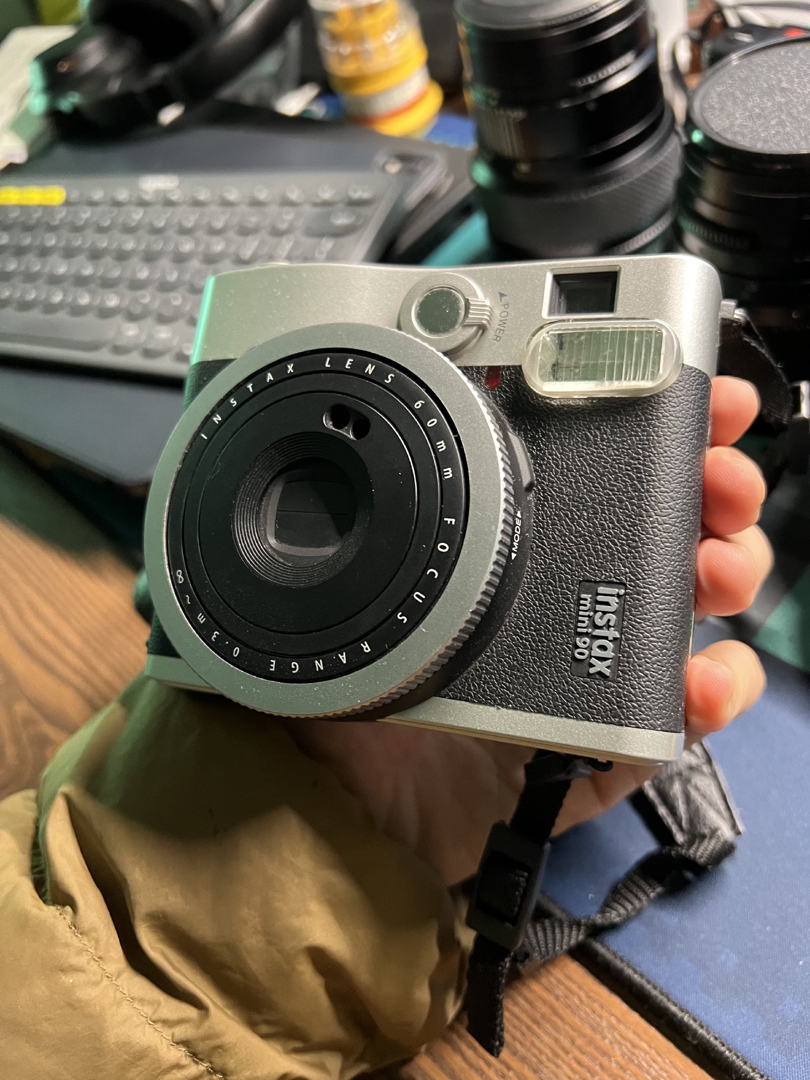 手里拿着一台银黑配色的 instax mini 90,背后是键盘和几颗镜头