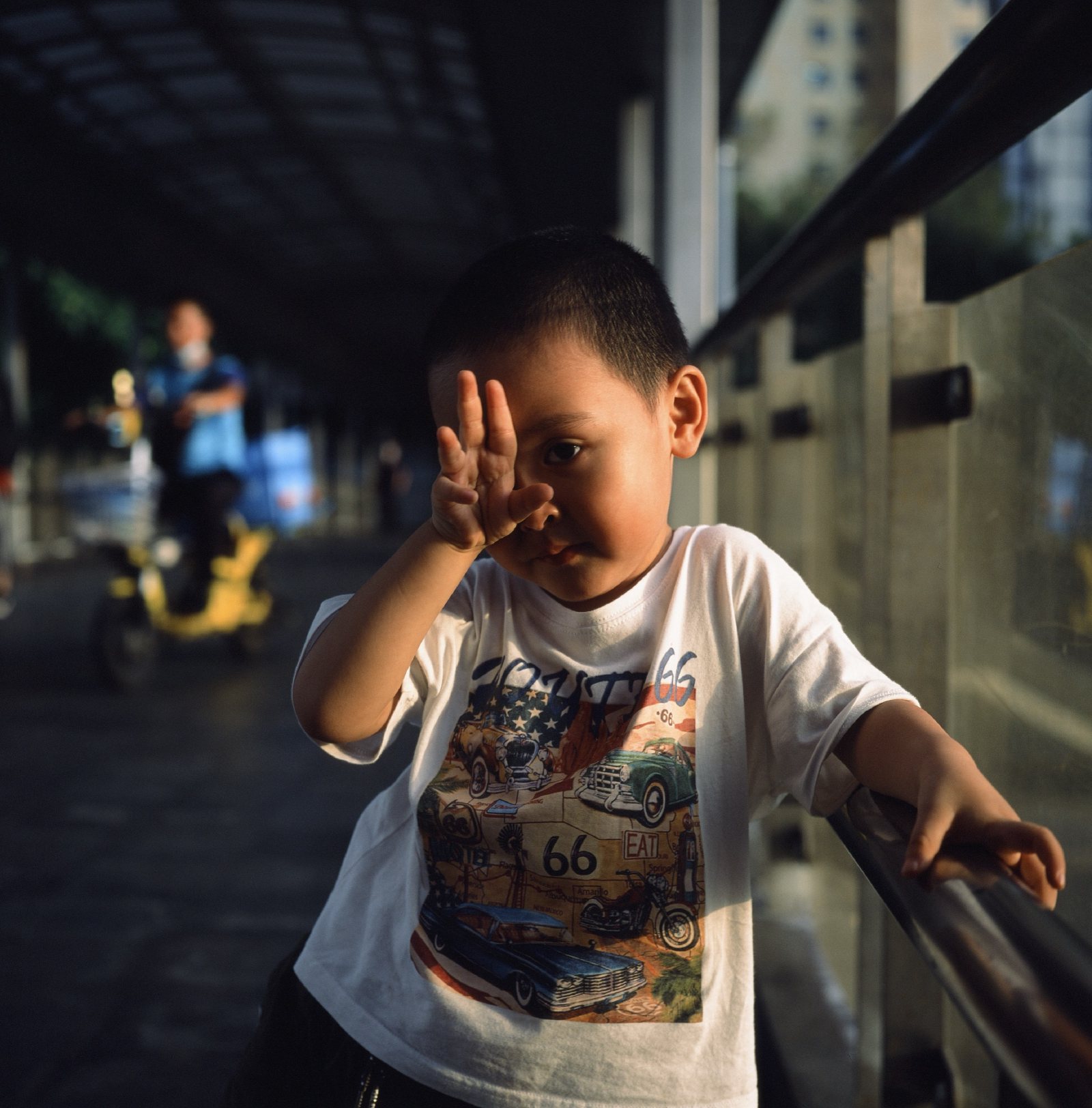 儿子举起一只手挡在脸前，背后有人骑电动车经过