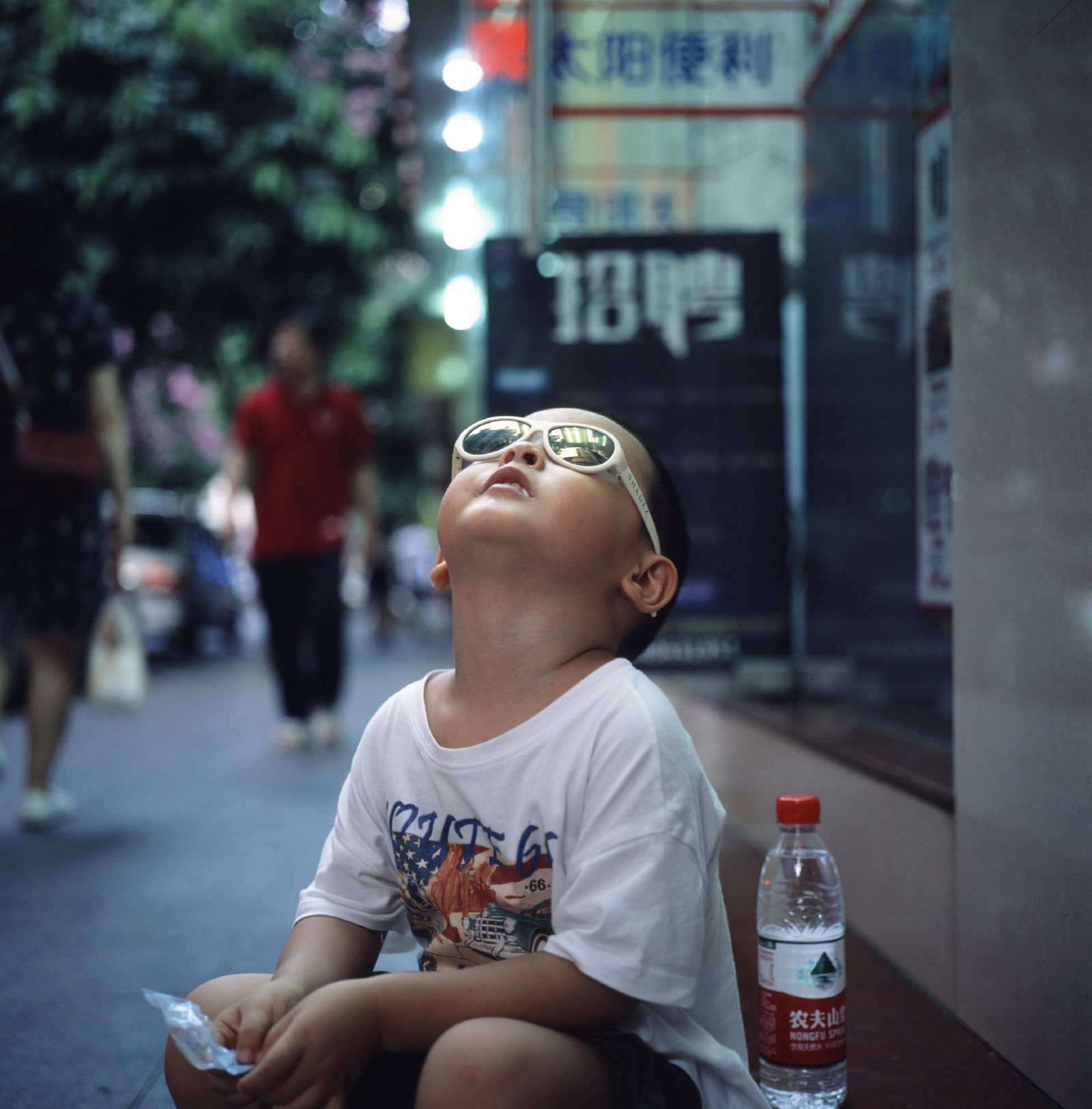 儿子戴着墨镜坐在店门口，仰头朝天，脚边一瓶矿泉水