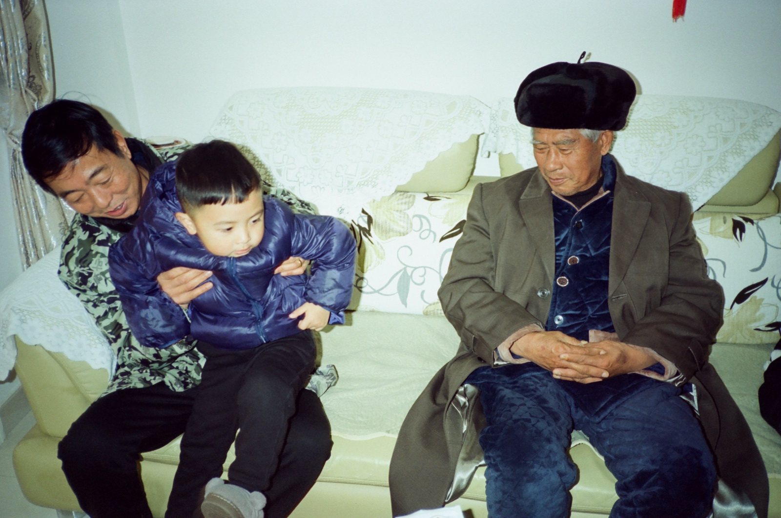 沙发上亲戚抱着儿子，旁边坐着戴帽子的长辈