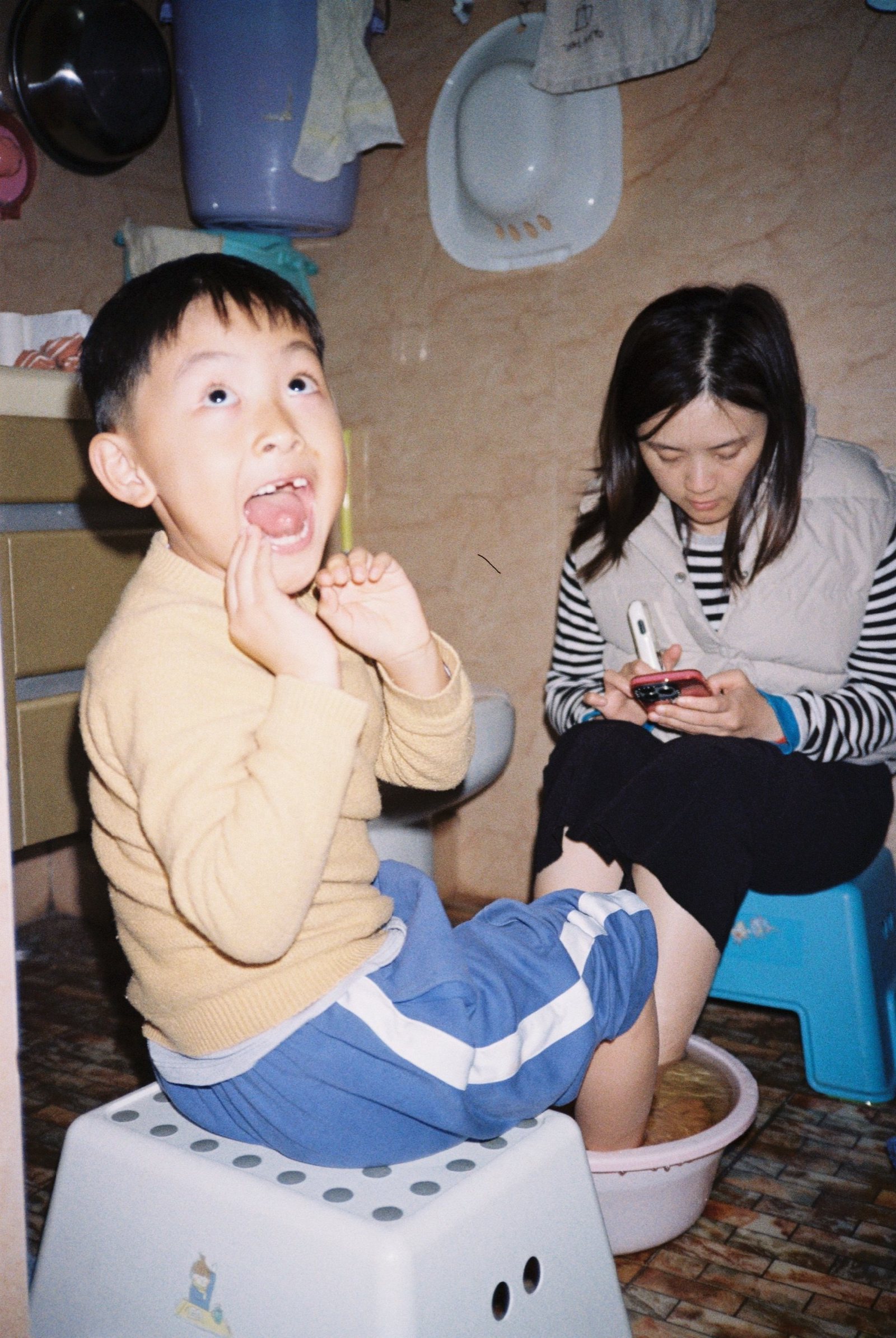 儿子坐在小凳上泡脚仰头，妈妈在旁边看手机